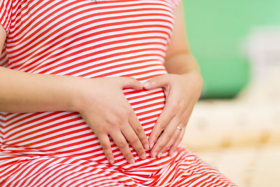 Midsection of pregnant woman making heart shape on abdomen indoor