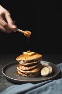 Close up view of stack of pancake with butter and banana in plate a hand is pouring honey on pancake