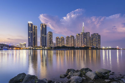 Illuminated modern buildings by bay against sky