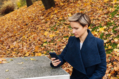 Short-haired blonde uses a smartphone sitting at a table in the park in the fall