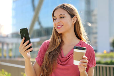 Smiling young woman using mobile while holding coffee