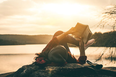 Woman reading book while lying on lakeshore during sunset