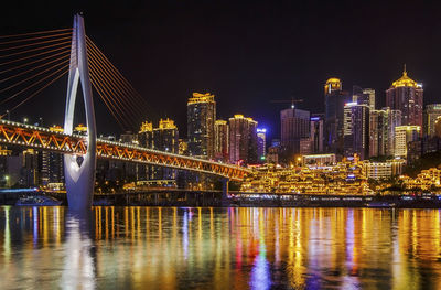 Illuminated bridge over river by buildings against sky at night