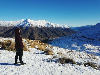 Side view of man wearing hood while standing on snowcapped mountains against clear blue sky