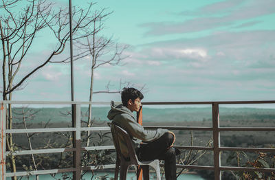 Full length of young man sitting on railing against sky