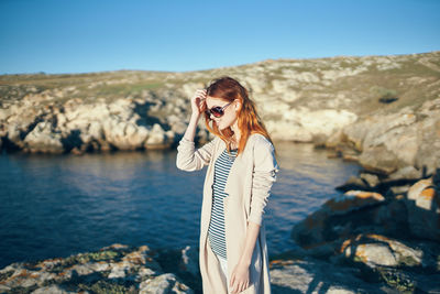 Young woman wearing sunglasses standing on rock against sky