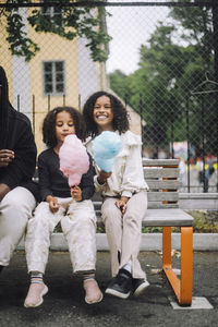 Happy sisters holding cotton candies while sitting on bench at amusement park