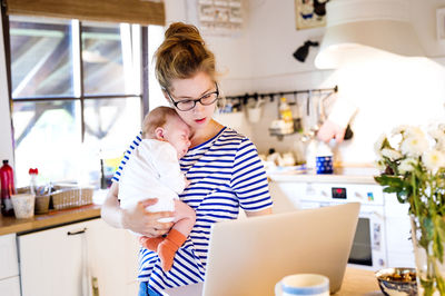 Mother with baby in kitchen looking at laptop