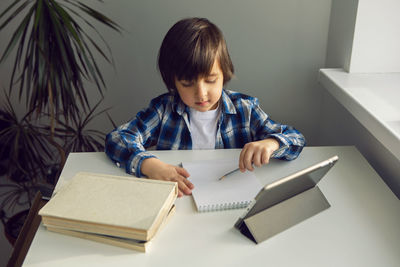 Boy child sits at a desk and learns with a tablet notebook and book