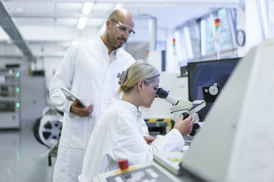 Male technician standing by female scientist looking into microscope while doing research in laboratory