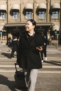 Smiling businesswoman with luggage holding smart phone while crossing street in city