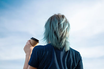 Unrecognisable blue haired woman drinks take away coffee against blue sky.