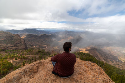 Rear view of man sitting on mountain against sky