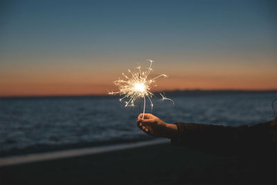 Person hand holding sparkler at sea against sky during sunset