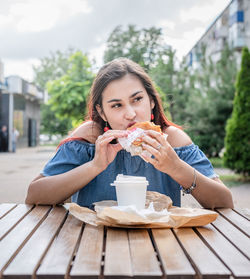 Hungry stylish woman, enjoying eating a burger outdoors, dressed in jeans shirt, wearing sunglasses