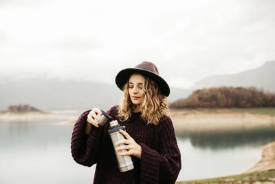 Young woman wearing hat standing against lake during winter