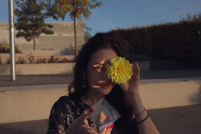 Portrait of young woman holding flower and prism while standing outdoors