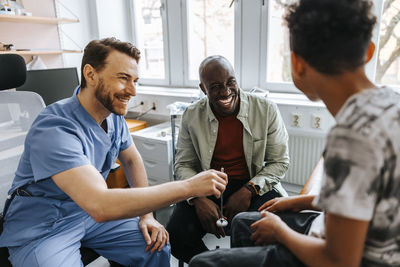 Smiling male healthcare worker holding reflex hammer while sitting with father and son during visit in examination room