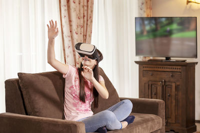 Teenage girl gesturing while using virtual reality simulator on sofa at home