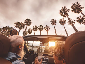 People photographing car on palm trees against sky during sunset