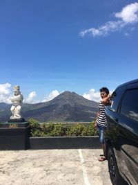 Young man standing by statue on mountain against blue sky