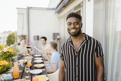 Portrait of smiling young man wearing striped shirt with friends enjoying dinner in balcony