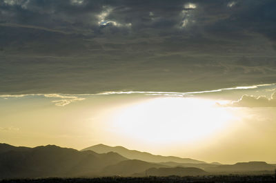 Mountain ranges, valleys, in silhouette under thick bank of rain clouds and a bright hazy sky