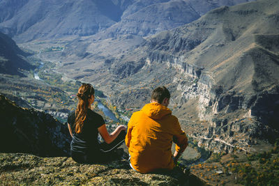 Rear view of couple sitting on cliff against mountains