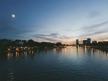 Illuminated buildings by river against sky during sunset