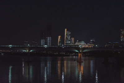 Illuminated bridge over river by buildings against sky at night