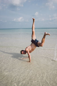 Full length of shirtless man practicing handstand in sea on shore at beach