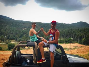 Full length of happy friends sitting on mountain against sky