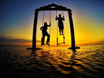 Silhouette people on swing at beach against sky during sunset