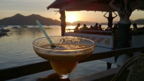 Hand holding bowl of soup with people in background at lakeside during sunset