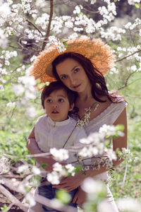 Woman in pink clothes and a straw hat stands with a child in white shirt in blossoming apple orchard