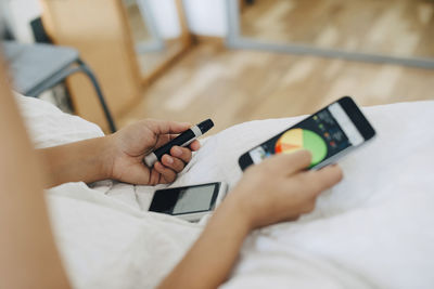 Cropped woman checking blood sugar level while using mobile phone on bed