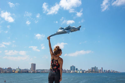 Rear view of woman waving shirt at riverbank against sky