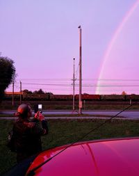 Rear view of man photographing rainbow over cars against sky