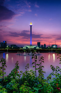 Illuminated buildings by river against sky at sunset