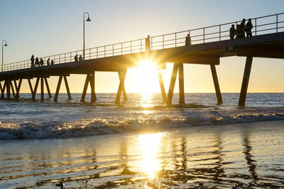 Low angle view of silhouette people walking on pier over sea at glenelg beach during sunset