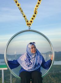 Woman looking away while sitting on chair swing against sky