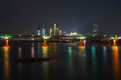 Illuminated bridge over river by buildings against sky at night