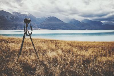 Camera on tripod over land by lake against sky