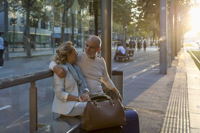 Spain, barcelona, senior couple with baggage sitting at tram stop in the city at sunset