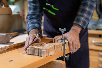 Crop anonymous male woodworker using metal clamp to fix carved wooden detail on workbench while filing with sand paper in carpentry workshop