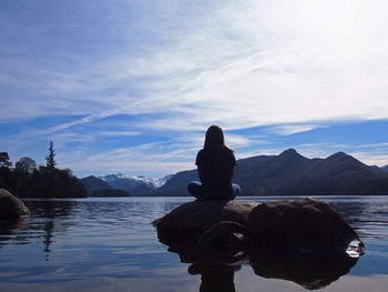 Rear view of young woman sitting on rock in lake against sky