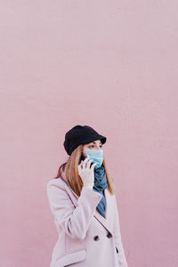 Woman wearing mask talking over smart phone while standing against wall