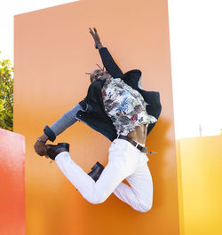Low angle of carefree african american male in fancy outfit in moment of jumping above ground near vibrant orange wall in city