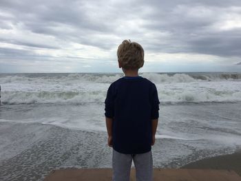 Rear view of man standing on beach against cloudy sky