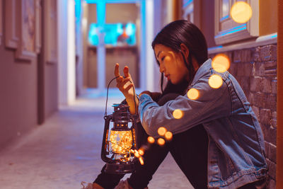 Side view of woman looking at illuminated lantern while sitting in corridor